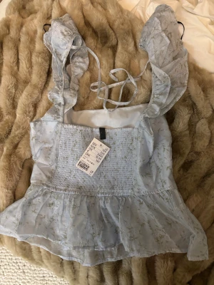 Blommig ljusblå blus från H&M Divided - Supersöt ljusblå blus med blommigt mönster från H&M Divided. Toppen har volangdetaljer vid axlarna, knytning i ryggen och smockad midja. Materialet är lätt och luftigt, perfekt för vår och sommar. Passformen är normal och toppen är ärmlös med breda volangaxelband.