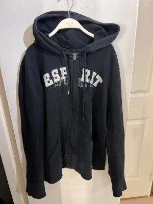Svart hoodie från Esprit med dragkedja - Svart hoodie från Esprit med vit text på bröstet. Tröjan har huva med snörning, långa ärmar och praktiska fickor framtill. Materialet är mjukt och skönt, perfekt för chill dagar. 