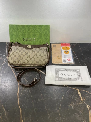 Gucci axelväska med GG-mönster - Snygg axelväska från Gucci med klassiskt GG-mönster i beige och brunt. Väskan har bruna skinn-detaljer, justerbar axelrem och guldfärgad GG-logga framtill. Stängs med dragkedja och har ett fack inuti. Perfekt storlek för det viktigaste.