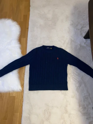 Marinblå stickad tröja Polo Ralph Lauren - Snygg marinblå stickad tröja från Polo Ralph Lauren i storlek S. Tröjan har klassiskt kabelstickat mönster, rund hals och långärmade ärmar. På bröstet finns den ikoniska röda Polo-loggan broderad. Perfekt för dig som gillar stilrena och tidlösa plagg. Köpte den när jag va utomlands och nu är den liten på mig och vill sälja den och i bra kvalitet den kommer liten jag är 158 och passar typ inte mig men kommer passar dem som är typ 145-150