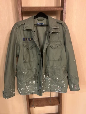 Raulp lauren field jacket - Ralph Lauren Field Jacket med Paint Splatter design Säljer en snygg och karaktärsfull Ralph Lauren field jacket i klassisk militärgrön färg. Jackan har en autentisk paint splatter finish som ger den ett unikt, vintage inspirerat uttryck. Perfekt för dig som gillar plagg med attityd och personlighet.