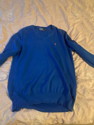 Blå v-ringad tröja från Polo Ralph Lauren - Snygg blå v-ringad tröja från Polo Ralph Lauren med klassisk röd logga broderad på bröstet. Tröjan har långa ärmar och ribbade muddar vid ärmslut och nederkant. Perfekt för dig som gillar stilren och enkel design.