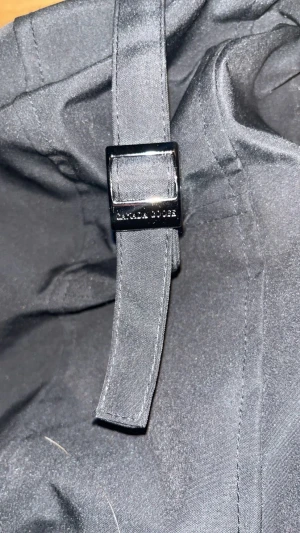 Svart pufferjacka från Axel Arigato - Säljer en svart pufferjacka från Axel Arigato med huva och snyggt spänne på kragen. Jackan har quiltad design, knappar framtill och en clean look som passar perfekt när det är kallt ute. Materialet är polyester och jackan har en loose passform.