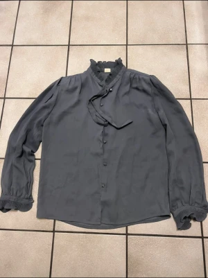 Mörkgrå blus med volangkrage storlek S/M - Snygg mörkgrå blus i mjukt, tunt tyg med volangdetaljer vid krage och ärmslut. Knäppning framtill med tygklädda knappar och dekorativt band vid halsen. Långa ärmar med puff och breda manschetter som har pärlknapp. Perfekt för dig som gillar en chic och vintageinspirerad look.