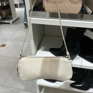 Beige axelväska med dragkedja - Snygg beige axelväska med stilren, avlång form och diskret mönstrad yta. Väskan har en smal axelrem och praktisk dragkedja upptill. Perfekt för att bära med sig det viktigaste och passar till många olika outfits.