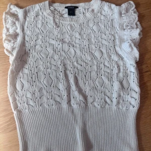 Vit stickad topp med volangärm  - Supersöt vit stickad topp från Lindex med volangärmar och snyggt hålmönster. Toppen är ärmlös med ribbad midja och rund halsringning. 