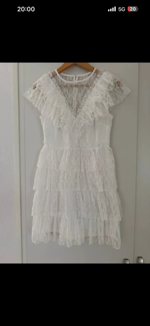 Vit spetsklänning från Bubbleroom - Supersöt vit spetsklänning från Bubbleroom med volanger och pärlknappar i ryggen. Kort modell med lager på lager-effekt och blommigt spetsmönster. Klänningen har korta ärmar och rund halsringning, perfekt för dig som gillar romantisk stil. Saknar en knapp vid ryggen bara med inget som syns!