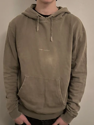 Olivgrön hoodie från Saint Laurent - Säljer en olivgrön hoodie från Saint Laurent Paris med broderad logga på bröstet. Det är beat down stil. Hoodien har klassisk känguruficka, ribbade muddar och justerbar huva med snören. Materialet är mjukt och bekvämt, perfekt för chill dagar. Snygg och enkel design som funkar till jeans eller joggers. Modellen på bilden är 186 och väger 70 kg, 