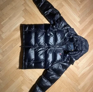 Svart/mörkblå pufferjacka Polo Ralph Lauren XL - Snygg svart pufferjacka från Polo Ralph Lauren i storlek XL. Jackan har glansig finish, huva och flera praktiska fickor med dragkedja. Perfekt för kalla dagar och har en klassisk, sportig look med diskret logga på bröstet.