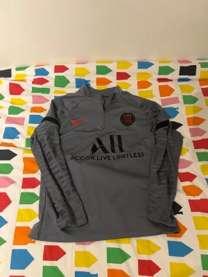 Grå PSG långärmad träningströja Nike - Säljer en grå långärmad träningströja från Nike med PSG-logga och Accor Live Limitless-tryck på bröstet. Tröjan har half zip, svarta detaljer och mönstrade ärmar. På ryggen står det 'Visit Rwanda'. Perfekt för fotbollsträning eller chill.