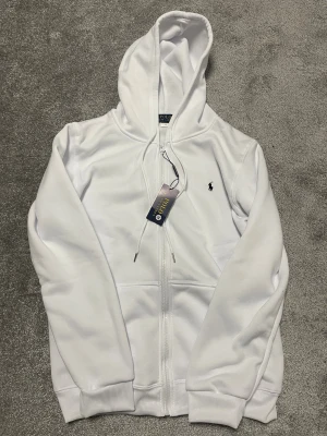 Vit hoodie från Polo Ralph Lauren - Vit hoodie från Polo Ralph Lauren med klassisk broderad logga på bröstet. Tröjan har huva med snörning, dragkedja framtill och två fickor. Materialet är mjukt och känns skönt mot huden. Perfekt för chill dagar och snygg streetstyle.