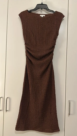 Brun strukturerad klänning från H&M - Säljer en brun kortklänning från H&M i storlek S. Klänningen har en snygg strukturerad yta med vågigt mönster och är ärmlös med en rak, enkel siluett. Perfekt för dig som gillar stilrena och trendiga plagg med det lilla extra.