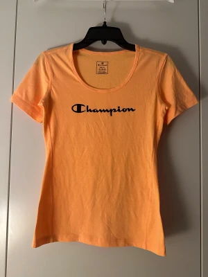 Orange t-shirt från Champion - Snygg orange t-shirt från Champion med svart logotyp tryckt på bröstet. Modellen är figurnära med rund hals och korta ärmar. Tillverkad i mjuk bomull som känns skön mot huden. Perfekt för dig som gillar sportiga och stilrena plagg.