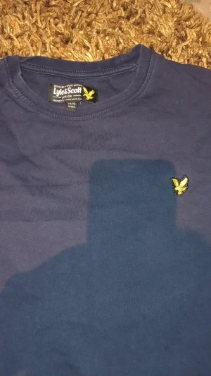 Marinblå t-shirt från Lyle & Scott - Snygg marinblå t-shirt från Lyle & Scott med klassisk rund hals och korta ärmar. På bröstet sitter den ikoniska gula örnloggan. Tillverkad i mjuk bomull som känns skön mot huden. Perfekt för dig som gillar stilrena och enkla plagg.