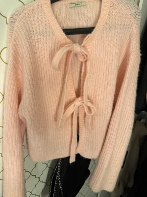 Ljusrosa stickad kofta från Gina Tricot - Superfin ljusrosa stickad kofta från Gina Tricot i storlek S. Koftan har två söta knytband framtill och är ribbstickad med långärmade ärmar. Perfekt för att ge din outfit en mjuk och cozy vibe.