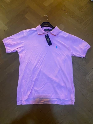  Polo pikétröja  - Snygg rosa pikétröja från Polo Ralph Lauren med klassisk krage och korta ärmar. Tröjan har den ikoniska blå broderade loggan på bröstet och är gjord i mjuk bomull. Perfekt för dig som gillar stilrena och färgglada plagg.