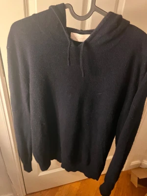 Mörkblå hoodie i karshmir från sayless - Snygg mörkblå hoodie från sayless i mjuk karshmir. Tröjan har huva med snörning och långa ärmar, perfekt för dig som gillar en chill och stilren look. Passformen är normal och materialet känns lyxigt och varmt. En klassisk hoodie som funkar året runt. Och är väldigt populär nästintill aldrig använd så ny skick.
