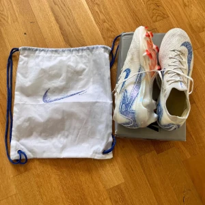 Nike mercurial vapor 16 - Säljer ett par Nike Mercurial vapor 16. Skorna är vita med blå detaljer och har en stickad ovandel samt snörning. Sulan är iriserande med orange Fg dobbar för bra grepp. Riktigt lätta och snygga skor för dig som vill sticka ut på planen. Ingen skopåse medföljer.