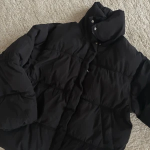 Svart pufferjacka från Divided - Svart pufferjacka från Divided med hög krage och dragkedja framtill. Jackan har en quiltad design och är tillverkad i syntetmaterial, perfekt för kalla dagar. Modellen är loose fit och har långa ärmar för extra värme.