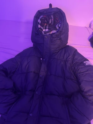 Burberry Jacka - Säljer en svart pufferjacka från Burberry med klassisk rutig insida i huvan. Jackan har huva med snörning, knappar och dragkedja framtill. Perfekt för kalla dagar och har en riktigt schysst oversized look. Storleken passar även Medium.