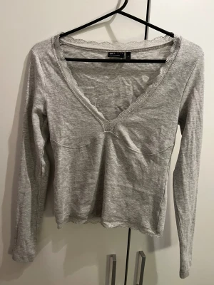 Grå långärmad topp med spets - Säljer en ljusgrå långärmad topp från ASOS i mjuk bomull. Toppen har v-ringad hals med söt spetskant och är figurnära i modellen. Perfekt för dig som gillar enkla detaljer och en feminin touch. Storlek S.
