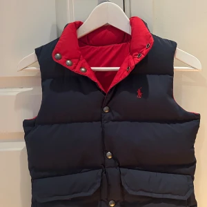 Mörkblå dunväst från Ralph Lauren - Snygg mörkblå dunväst från Ralph Lauren med röd insida och krage. Västen har tryckknappar med logga, två stora fickor framtill och klassisk broderad logga på bröstet. Perfekt för lager-på-lager och chill stil. Passar om man är 140-150 cm. Knappt använd. Ordinarie pris 1800kr. Diskuterar pris. 