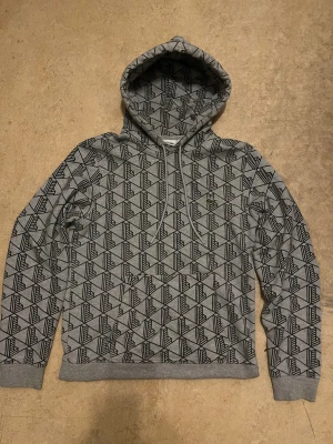 Lacoste monogram hoodie - En riktigt fet grå lacoste monogram hoodie. Den har ett unikt mönster och inga  fel på sig. Köpt för 1500 säljs för 900