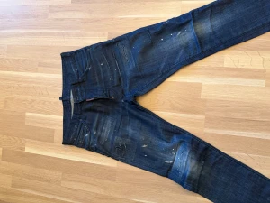 Mörkblå Dsquared2 jeans med slitningar - Snygga mörkblå jeans från Dsquared2 i storlek 48. Jeansen har coola slitningar, färgstänk och en tvättad look som ger dem en edgy vibe. Klassisk femficksmodell med Dsquared2-logga på bakfickan. Perfekta för dig som gillar streetstyle.