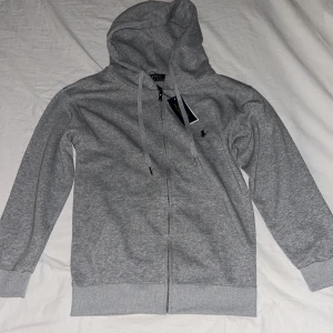 Grå Polo Ralph Lauren hoodie - Snygg grå hoodie från Polo Ralph Lauren med dragkedja och huva. Tröjan har långa ärmar, snörning vid huvan och klassisk Polo-logga broderad på bröstet. Tillverkad i mjuk bomull för skön komfort. Perfekt för chill dagar och streetwear. Storlek S men lite kortare armar