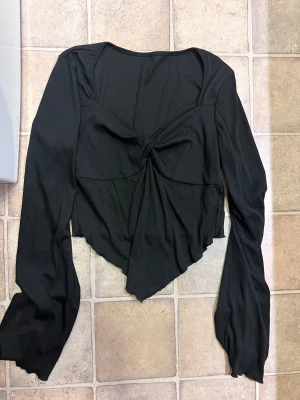 Svart långärmad topp med knut - Svart långärmad topp med snygg knutdetalj framtill och asymmetrisk nederkant. Toppen har en croppad passform och är tillverkad i ett mjukt, stretchigt material. Perfekt för dig som gillar trendiga och unika plagg.