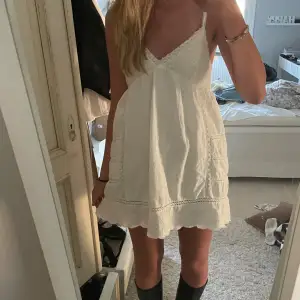 Supersöt vit kort klänning med tunna axelband och v-ringad hals. Klänningen har broderade detaljer och spets längs kanterna, samt en knytning i ryggen. Tillverkad i mjuk bomull, perfekt för varma dagar. Funkar även perfekt till studenten!