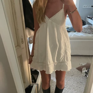 Vit kort sommarklänning med spets - Supersöt vit kort klänning med tunna axelband och v-ringad hals. Klänningen har broderade detaljer och spets längs kanterna, samt en knytning i ryggen. Tillverkad i mjuk bomull, perfekt för varma dagar. Funkar även perfekt till studenten!