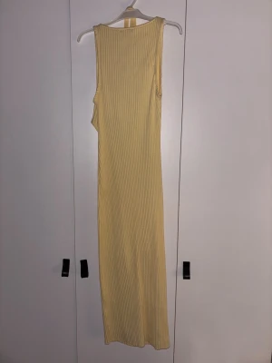 Ribbad ärmlös långklänning i gul - Snygg ribbad långklänning i ljusgul färg med ärmlös design och rund hals. Klänningen har en smickrande rak passform och är tillverkad i ett mjukt, stretchigt material som sitter bekvämt mot kroppen. Perfekt för dig som gillar minimalistisk och stilren look. Knyter i ryggen.