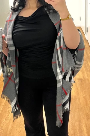 Grå rutig halsduk från Burberry - Snygg grå halsduk från Burberry med klassiskt rutmönster i svart, vitt och rött. Halsduken har fransade kanter och broderad Burberry-logga i gult. Perfekt accessoar för att lyfta din outfit med en lyxig touch.