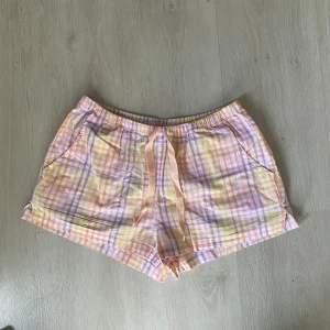 Pastellrutiga shorts med snörning - Säljer ett par mjuka shorts med pastellrutig design i lila, rosa, gul, orange och vitt. De har elastisk midja med snörning framtill och två sidofickor. Perfekta för chill dagar och sommarhäng. Materialet är lätt bomull som känns skönt mot huden.