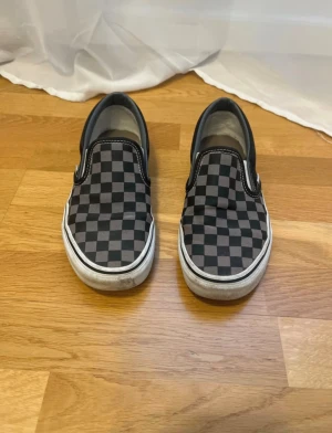 Vans slip-on checkerboard sneakers - Klassiska Vans slip-on sneakers med svart och grått rutigt checkerboard-mönster. Skorna har vit sula, svarta detaljer och är tillverkade i canvas med elastiska sidor för enkel påtagning. Perfekt för en avslappnad och cool streetstil. Sparsamt använda.