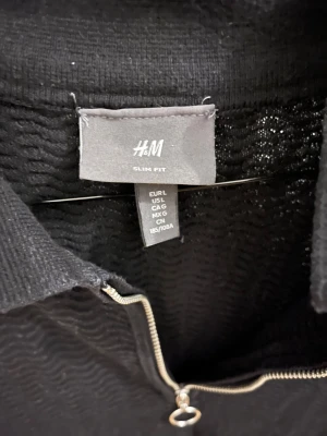 Svart pikétröja med dragkedja H&M - Svart pikétröja från H&M i slim fit med korta ärmar och krage. Tröjan har en snygg silverfärgad dragkedja med ringdetalj framtill. Materialet är stickat och har en vågig struktur som ger en cool känsla. Perfekt för dig som gillar stilrena och moderna plagg.