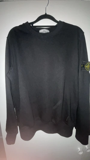 Svart sweatshirt från Stone Island - Svart långärmad sweatshirt från Stone Island med klassisk rund hals och ikonisk patch på vänster ärm. Tröjan är tillverkad i mjuk bomull och har ribbade muddar vid ärmslut och nederkant. Perfekt för dig som gillar stilrena och snygga streetwear-plagg.finns påse till och pris kan diskuteras 