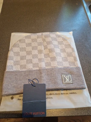 Grå rutig Louis Vuitton mössa - Snygg grå mössa från Louis Vuitton med klassiskt rutigt mönster och LV-logga framtill. Mössan har en bred uppvikt kant och är tillverkad i mjukt material som känns skönt mot huvudet. Perfekt för dig som gillar stilrena accessoarer.