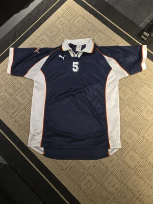 Blå Puma t-shirt med nummer 5 XXL - Sportig t-shirt från Puma i marinblått med vita och orange detaljer, samt stort nummer 5 på både fram- och baksida. Klassisk krage och korta ärmar, tillverkad i glansig polyester som ger en cool vibe. Perfekt för dig som gillar retro sportstil.