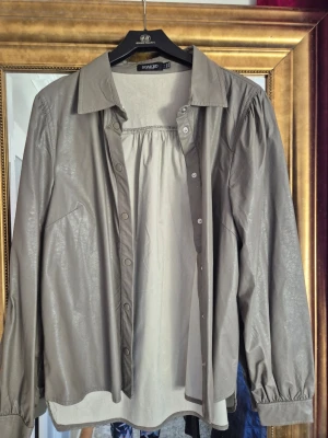 Grå skjorta i skinnlook från Soaked - Snygg grå skjorta från Soaked in Luxury med skinnliknande yta och klassisk krage. Långärmad modell med knappar framtill och lätt puffade ärmar. Perfekt för dig som gillar en cool och modern stil.