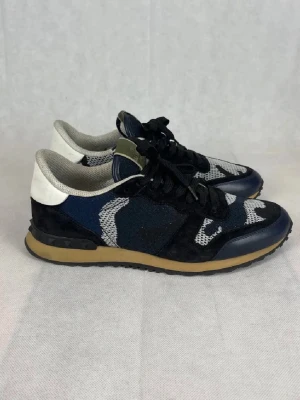 Valentino Rockrunner sneakers blå - Snygga Valentino Rockrunner sneakers med blå, svart och vita camouflage-detaljer. Skorna har snörning, rund tå och platt sula. Ovandelen är i en mix av mocka och textil, med beige inslag på sulan och vita partier vid hälen. Perfekt för dig som gillar exklusiva och unika sneakers.