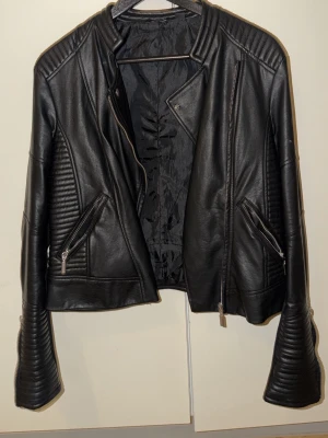 Svart skinnjacka med dragkedjor - Snygg svart skinnjacka med coola detaljer som quiltade partier på ärmar och sidor. Jackan har flera dragkedjor och en modern, lite bikerinspirerad look. Perfekt för dig som vill ha en edgy stil och gillar att sticka ut.