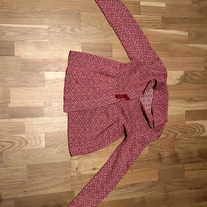 Rödvit mönstrad kofta från Mywear - Säljer en röd och vit kofta från Mywear med snyggt geometriskt mönster. Koftan är långärmad och har en klassisk passform. Perfekt för dig som gillar stickade plagg med lite retrovibe.