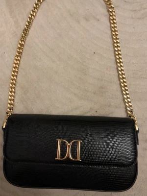 Svart axelväska från Dolce & Gabbana - Snygg svart axelväska från Dolce & Gabbana med guldfärgad kedja och logga framtill. Väskan är tillverkad i skinn med en elegant struktur och har ett rymligt fack på insidan. Perfekt för dig som vill ha en stilren och lyxig accessoar.