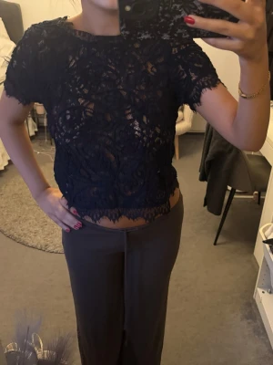 Mörkblå spetsblus med kort ärm - Säljer en mörkblå blus i spets med korta ärmar och rund hals. Blusen är croppad och har ett genombrutet mönster med dekorativ kant nedtill. 