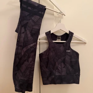 Svart träningsset från Stronger - Snyggt träningsset från Stronger med svart och mörkgrått bladliknande mönster. Setet består av ett sportigt linne och matchande tights i stretchigt material, perfekt för gymmet eller löprundan.