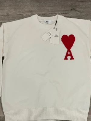 Ami paris - Säljer en vit stickad tröja från Ami Alexandre Mattiussi med röd hjärt- och A-logga på bröstet. Tröjan har rund hals, ribbade muddar och är långärmad. Perfekt för dig som gillar stilrena detaljer och vill ha något som sticker ut.
