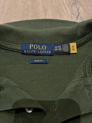 Grön pikétröja från Polo Ralph Lauren - Säljer en mörkgrön pikétröja från Polo Ralph Lauren i slim fit-modell. Tröjan har klassisk krage, korta ärmar och två knappar framtill. På bröstet finns den ikoniska gula Polo-loggan broderad. Materialet är mjuk bomull som känns skönt mot huden. XL i barn men Small/ Xs i vuxen 