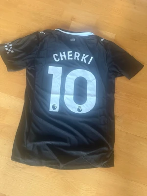 Svart Manchester City fotbollströja - Säljer en svart Manchester City matchtröja med CHERKI och nummer 10 på ryggen. Tröjan har vita detaljer, Premier League-logga och Etihad Airways tryck på framsidan. Materialet är lätt polyester och designen är modern med rund hals.